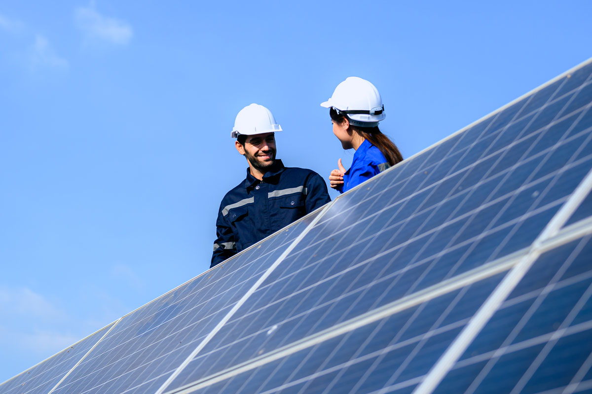 Zwei Personen mit Schutzhelmen arbeiten an Solarmodulen auf einer Dachfläche.